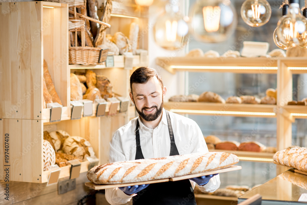 Handsome seller in uniform holding a big loaf of bread in the store with bakery products