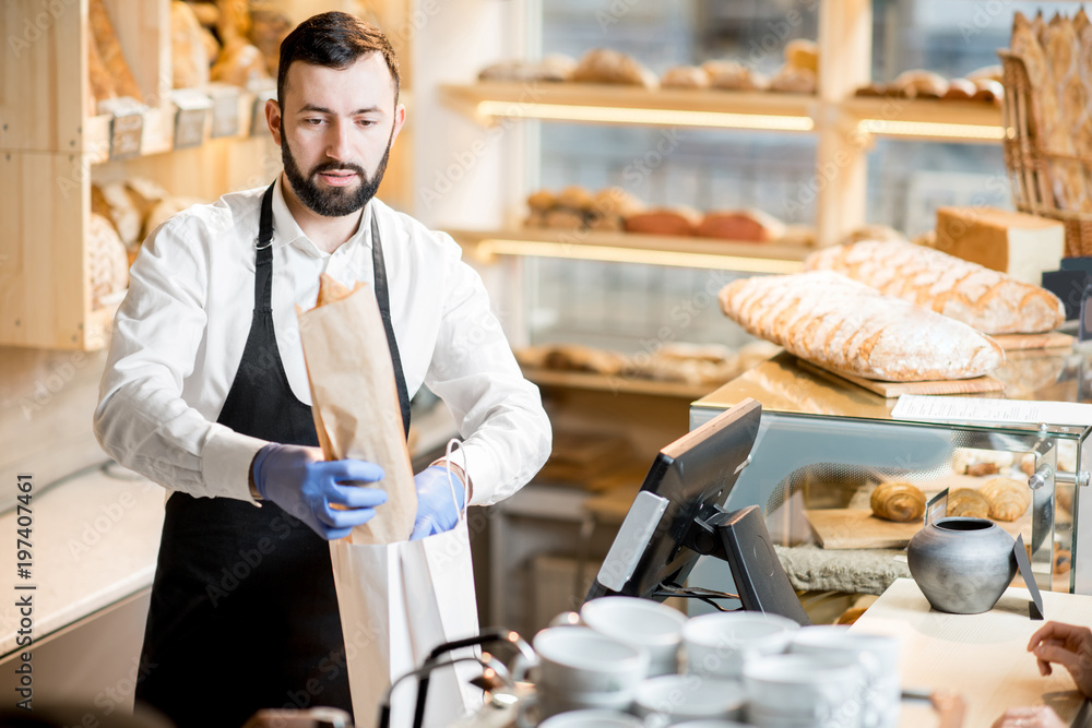 Bread seller packing a baguette into the paper bag in the store with bakery products