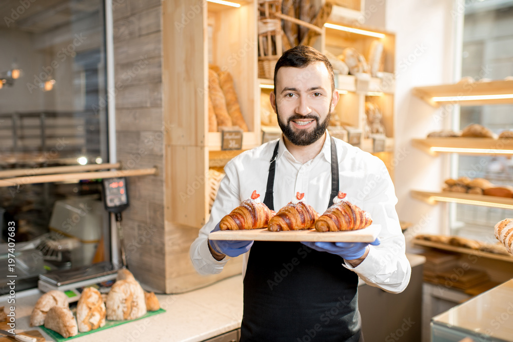 Portrait of a handsome seller in uniform standing with delicious croissants in the store with bakery