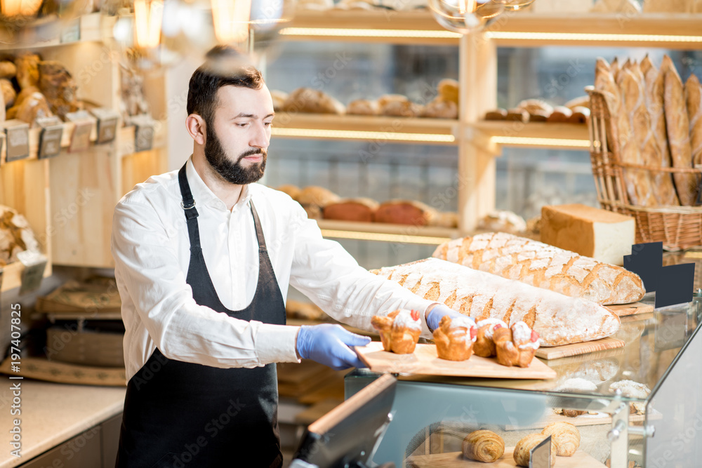 Bread seller putting maffins on the showcase standing in the beautiful store with bakery products