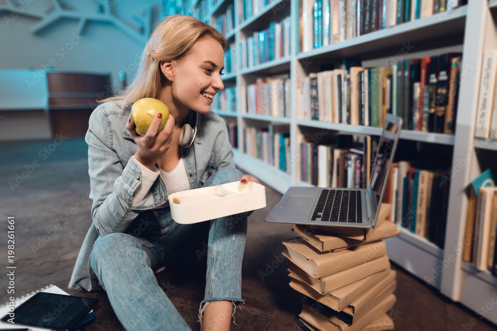 White girl near bookshelf in library. Student is eating apple from lunchbox.