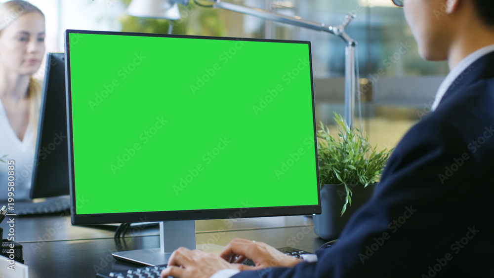 In the Office Businessman works at His Desk on a Personal Computer with Mock-up Green Screen. His Co