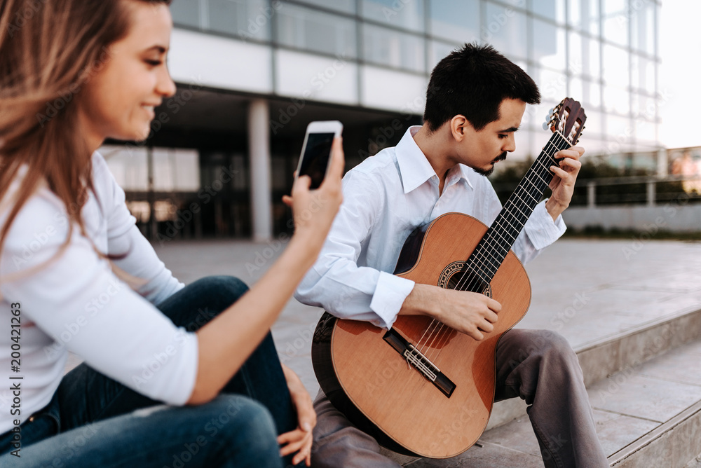 Young musician playing acoustic guitar in the city while being recorded on a phone by a girl.