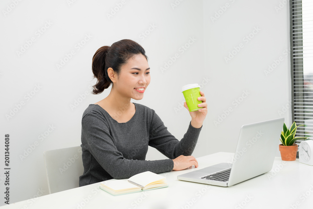 Young Asian female entrepreneur sitting at a table in her home office drinking a takeaway coffee and