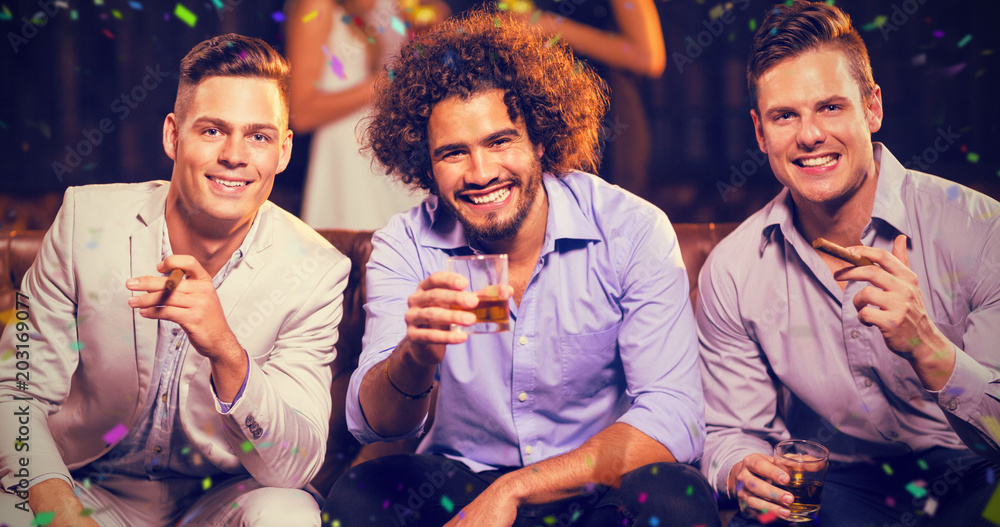 Three happy friends having cigar and whisky in bar against flying colours