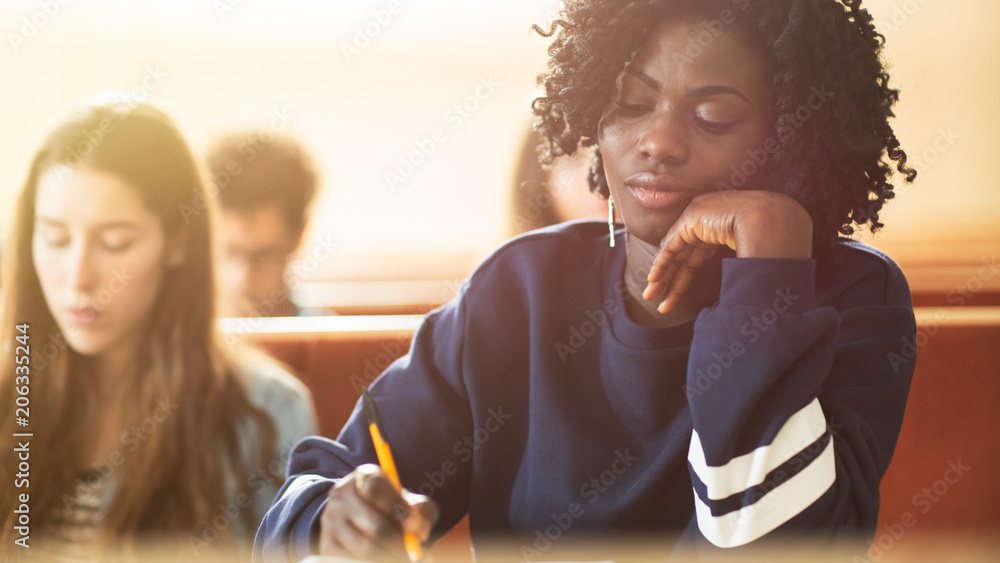 疲惫的黑人女学生坐在教室里的同学中间，她在教室里写作