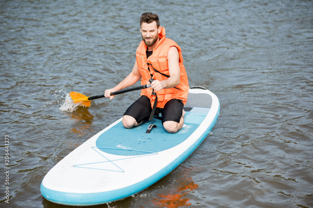 Man in life vest learning to swim with oar on the standup paddleboard on the lake