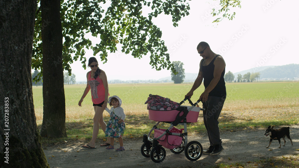 Cute young family of four go for a walk in the sunny park with their small dog