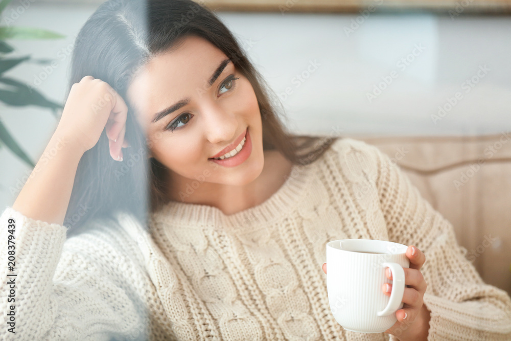 美丽的年轻女子在家里喝着一杯热茶，透过窗户观看