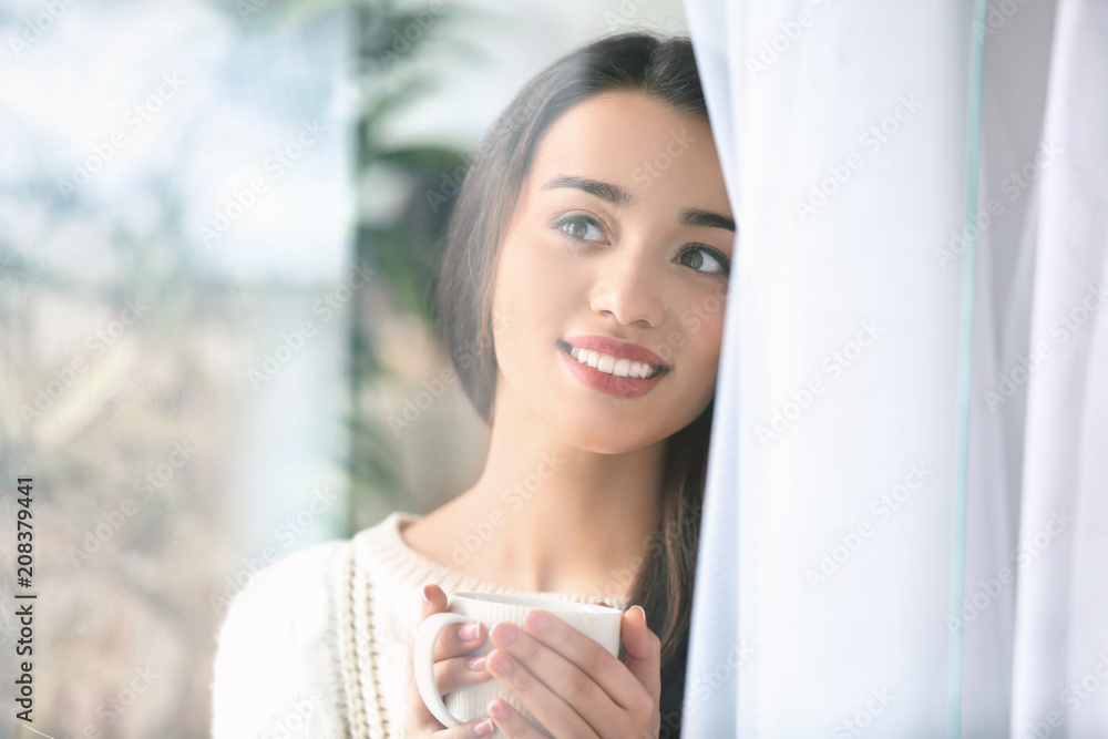 美丽的年轻女子在家里喝着一杯热茶，透过窗户观看