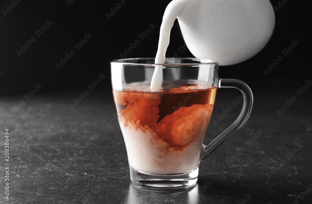 Pouring of milk into cup with tea on table against dark background