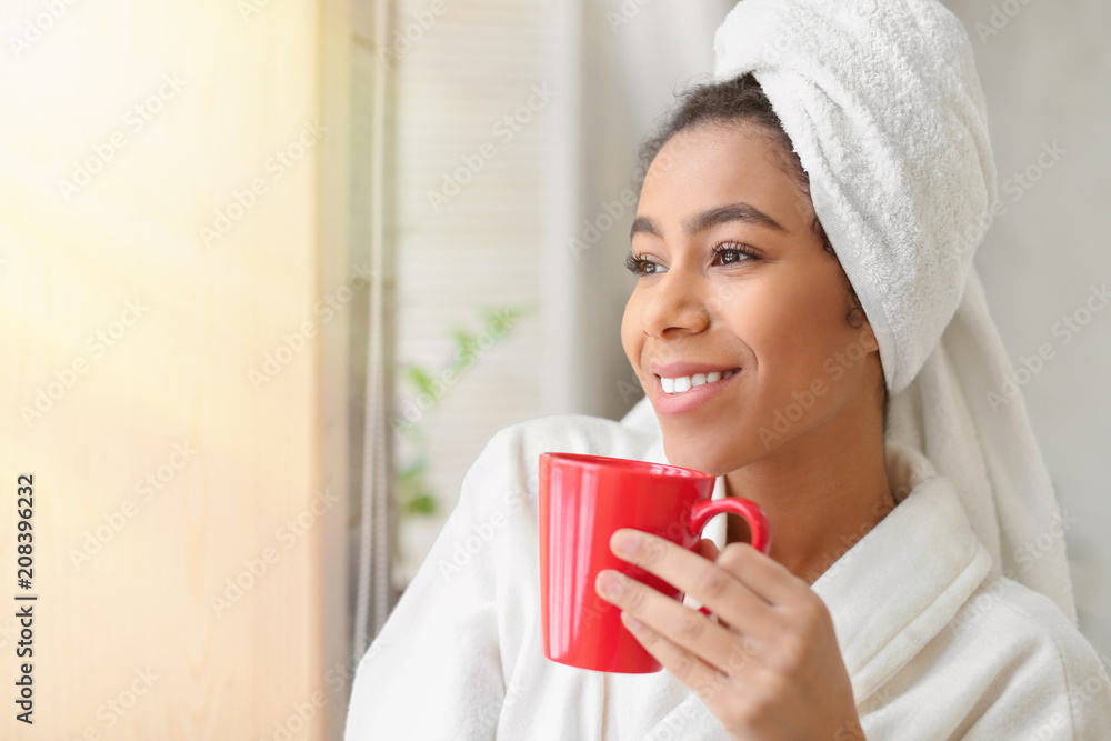 Beautiful African-American woman in bathrobe drinking tea at home