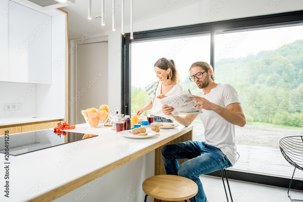 Young couple having breakfast cooking some food and reading newspaper at the kitchen of the modern c