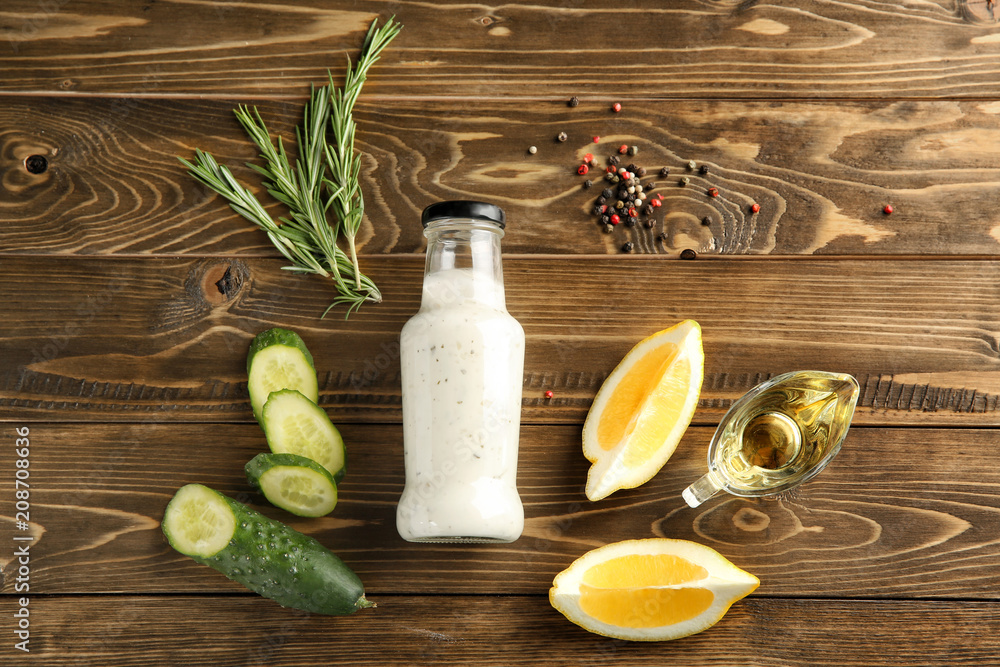 Bottle with tasty white sauce, cucumber and lemon slices on wooden background, flat lay