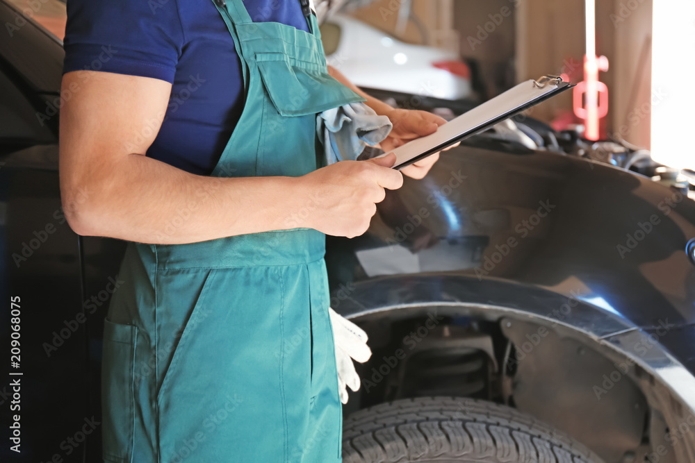 Young auto mechanic with clipboard near car in service center