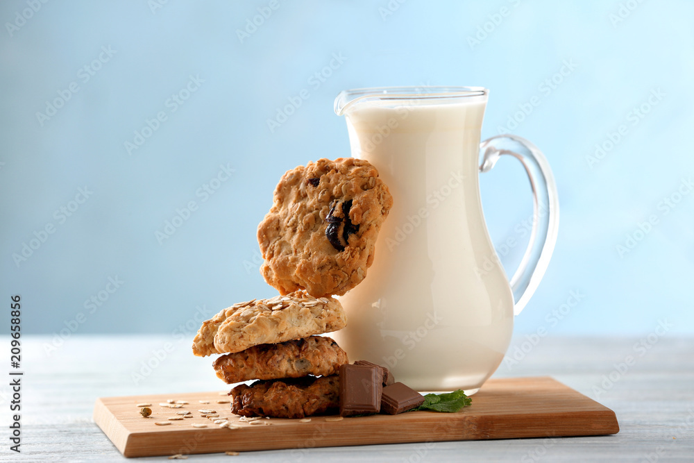 Wooden board with oatmeal cookies and jug of milk on table