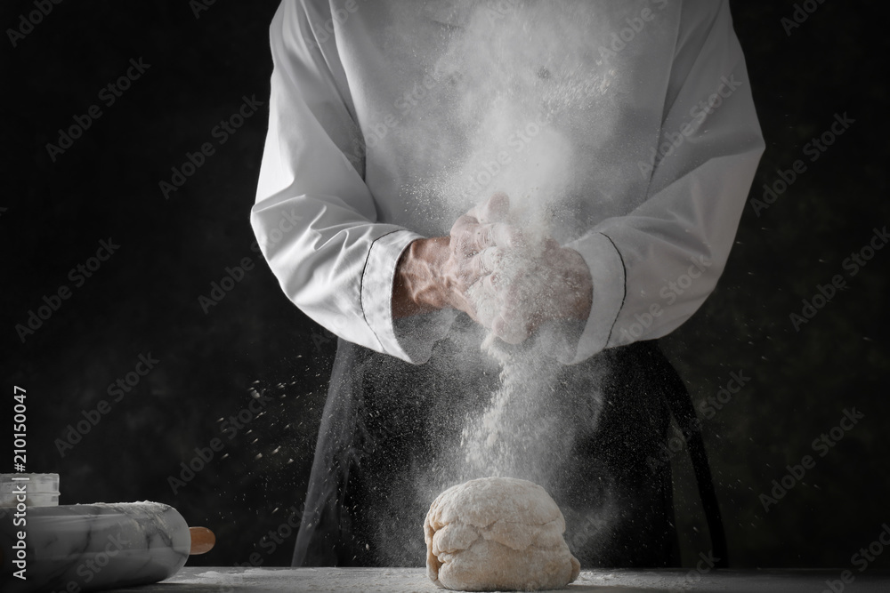 Man clapping and sprinkling flour over dough on dark background