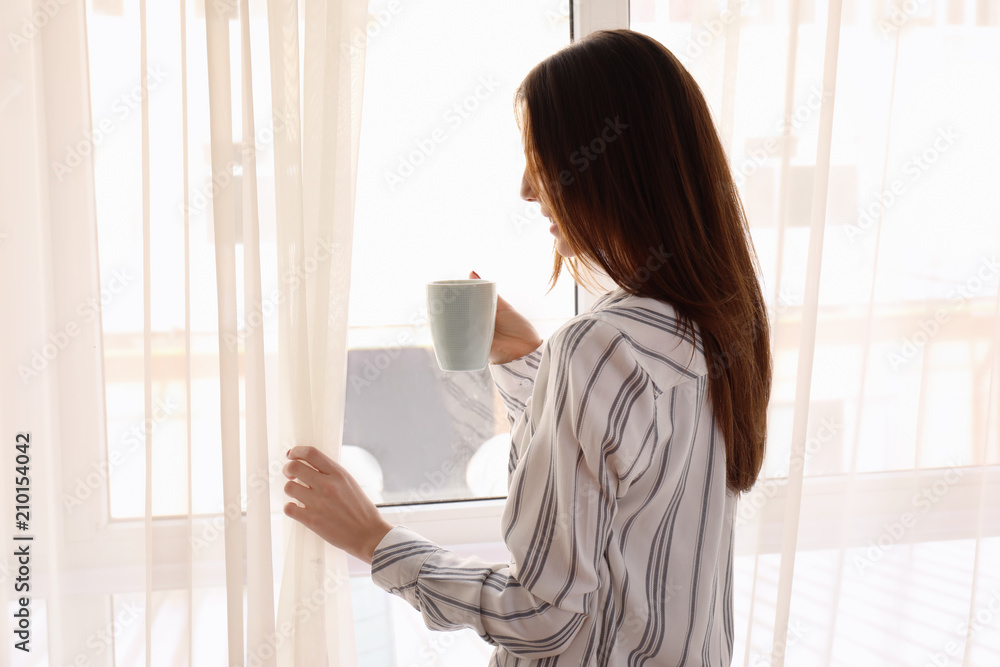 美丽年轻女子的早晨，在窗户附近喝着一杯咖啡