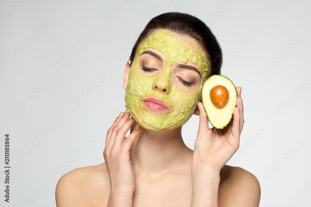 Beautiful young woman with facial mask and fresh avocado on light background