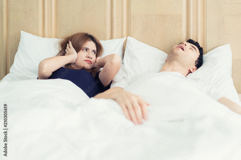 Annoyed Asian wife blocking her ears from noise of husband snoring in bedroom at home. Young couple 