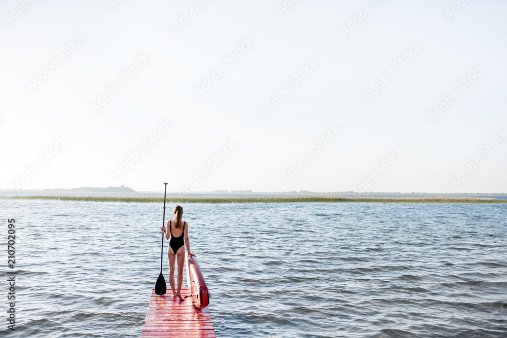 Beautiful woman standing back on the pier with paddleboard and oar enjoying morning view on the lake