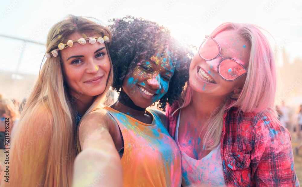 Multiethnic girls covered in colorful powder taking selfie at summer holi festival