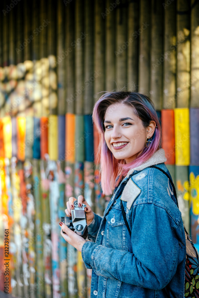 一位留着粉色头发、背着bakcpack的年轻女子在户外拿着相机。