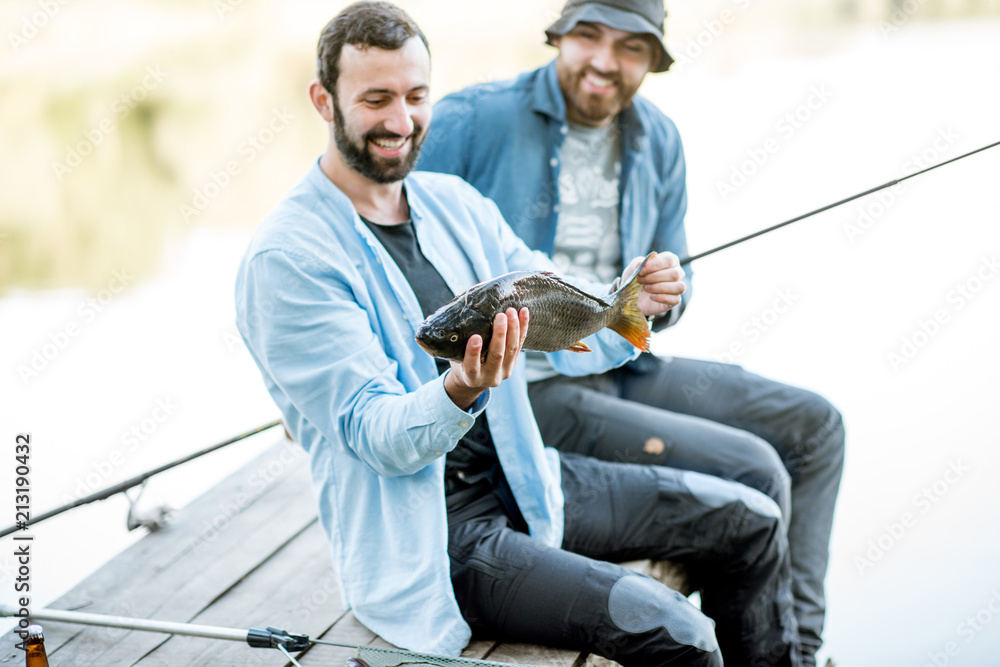 Two happy fishermen holding caught fish sitting on the wooden pier during the fishing on the lake at