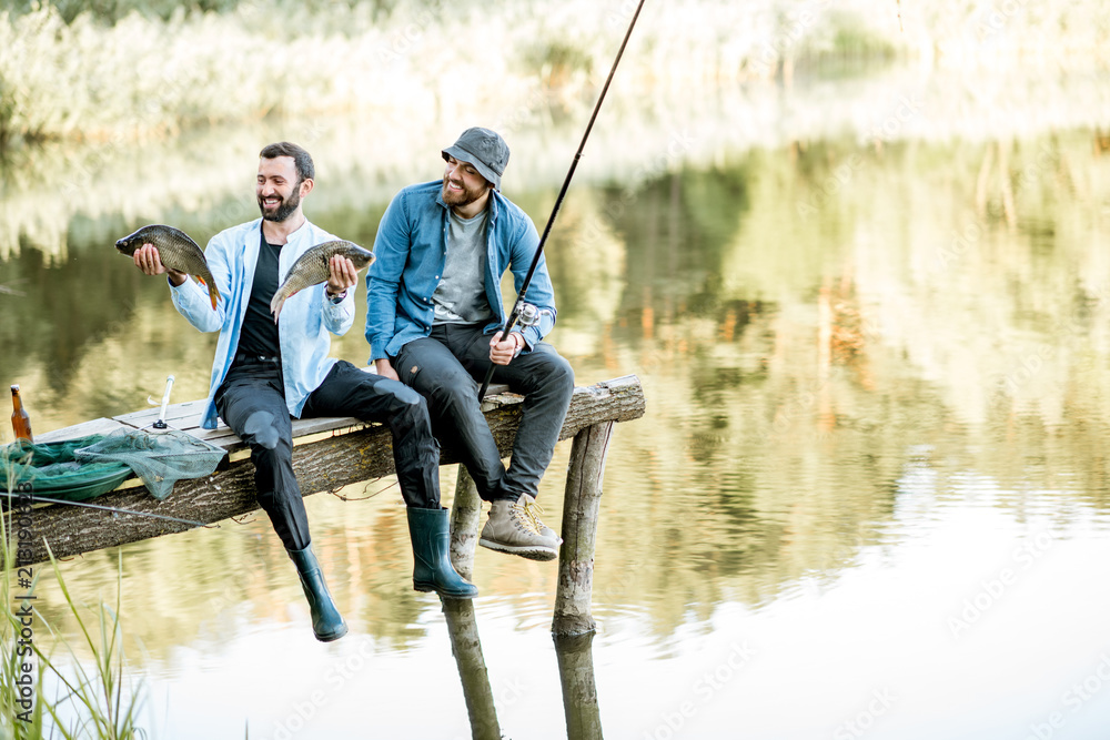 Two happy fishermen holding caught fish sitting on the wooden pier during the fishing on the lake at