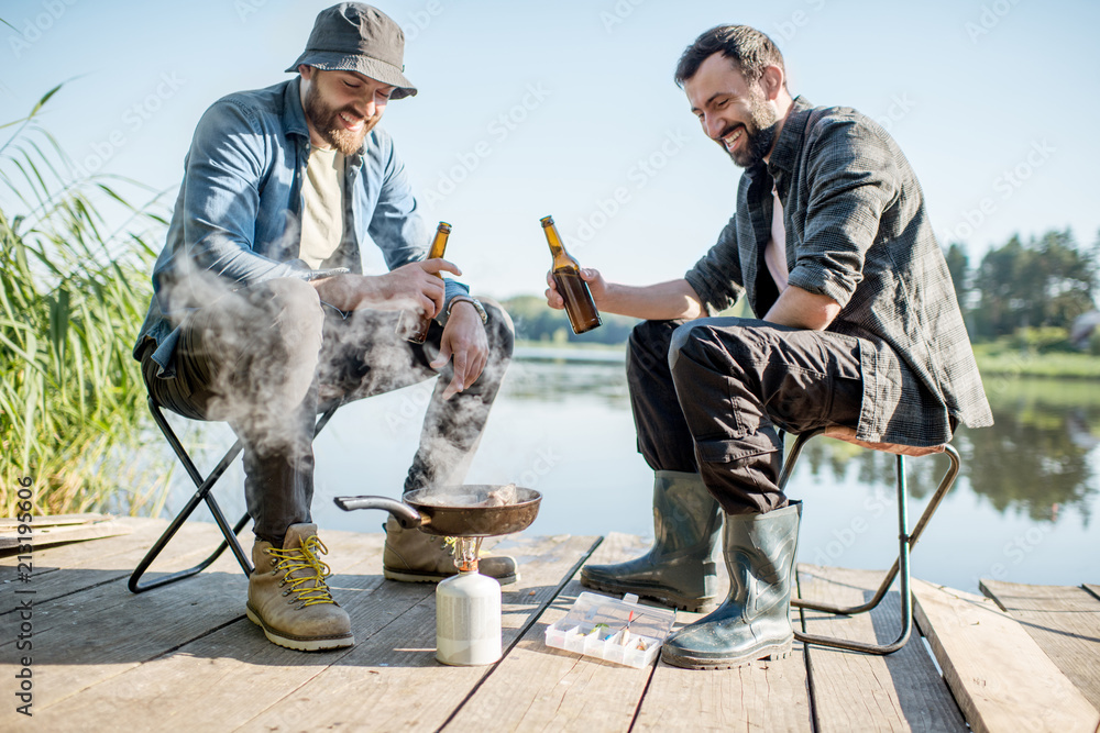 Two fishermen frying fish sitting with beer during the picnic on the wooden pier near the lake in th