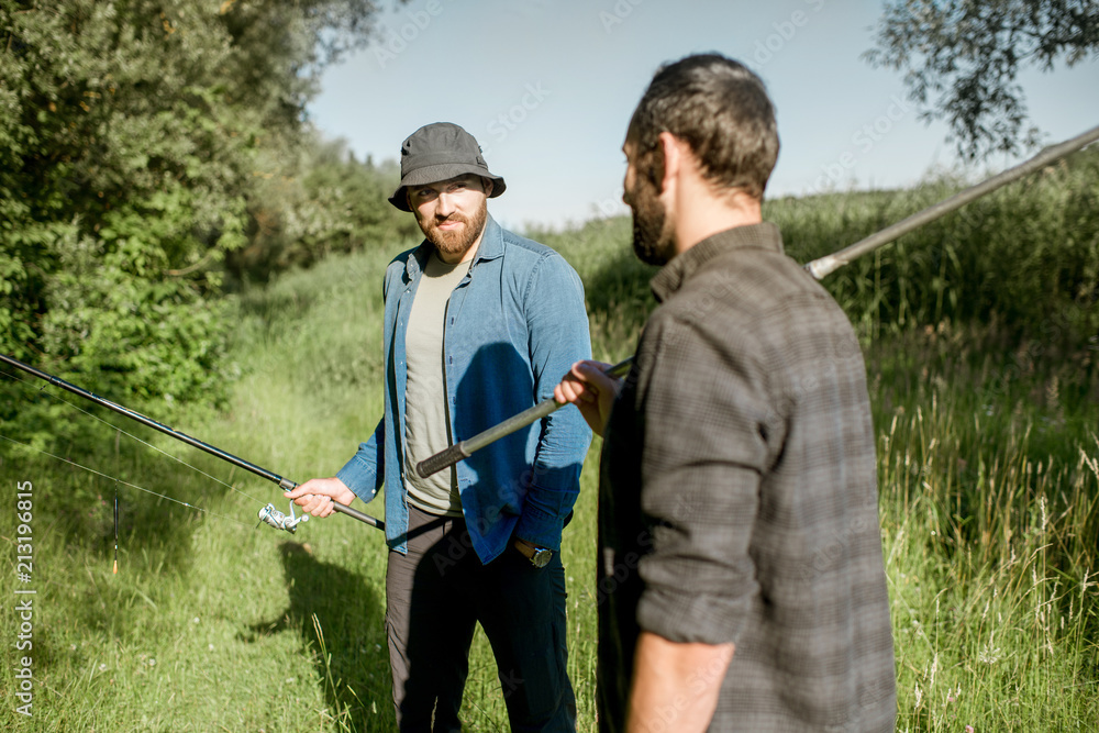 Two fishermen walking with fishing rod and net on the green lawn near the lake in the morning