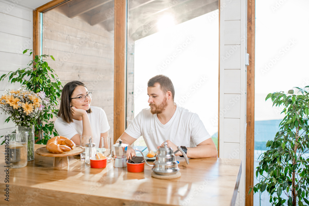 Young lovely couple in white t-shirts having a breakfast sitting at the table in the wooden house