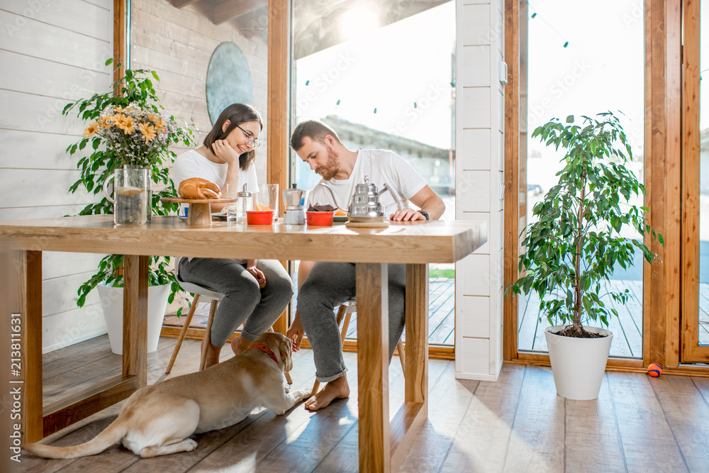 Young couple having a breakfast sitting at the table with their dog in the wooden country house