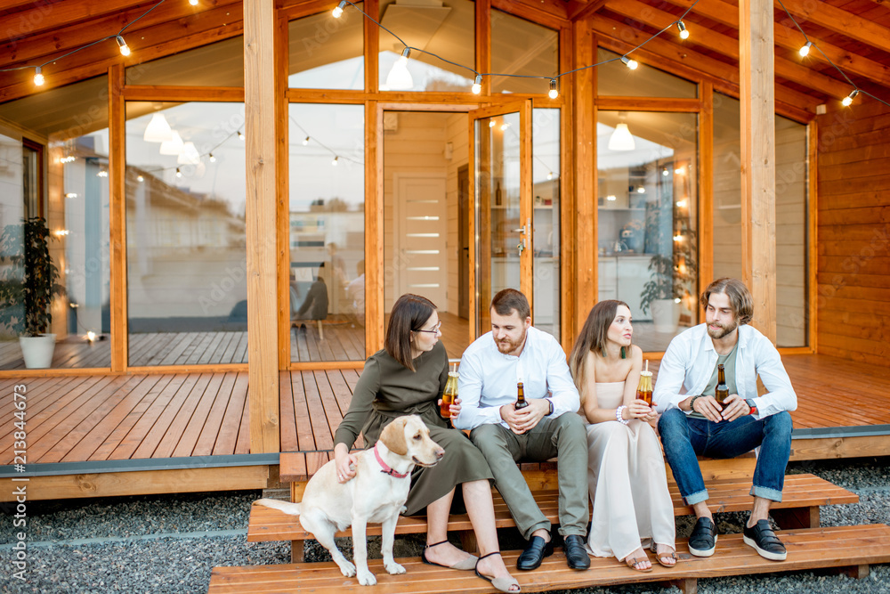 Young friends enjoying evening time sitting together with dog on the terrace of the modern wooden ho