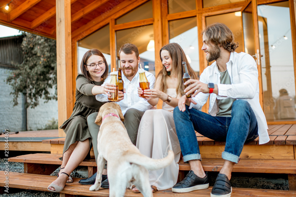 Young friends enjoying evening time sitting together with dog on the terrace of the modern wooden ho