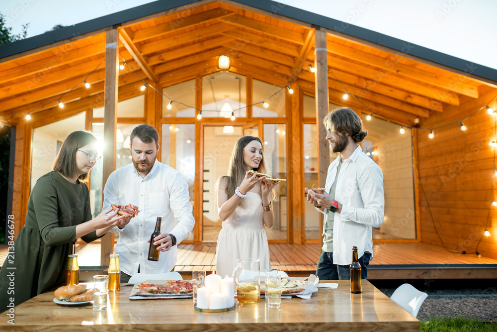 Friends having a dinner with pizza on the backyard of the modern house decorated with lights during 