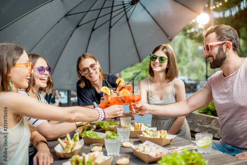 Young friends having fun together clinking glasses during the festive lunch outdoors in the park caf