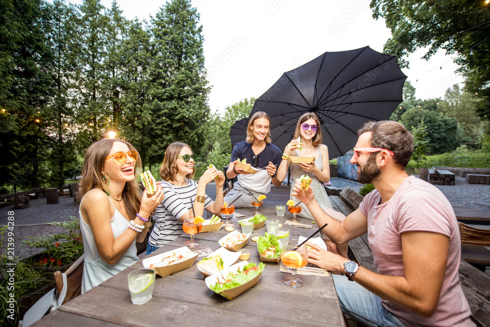 Young friends having fun sitting together with tasty snacks and drinks during the evening lights at 