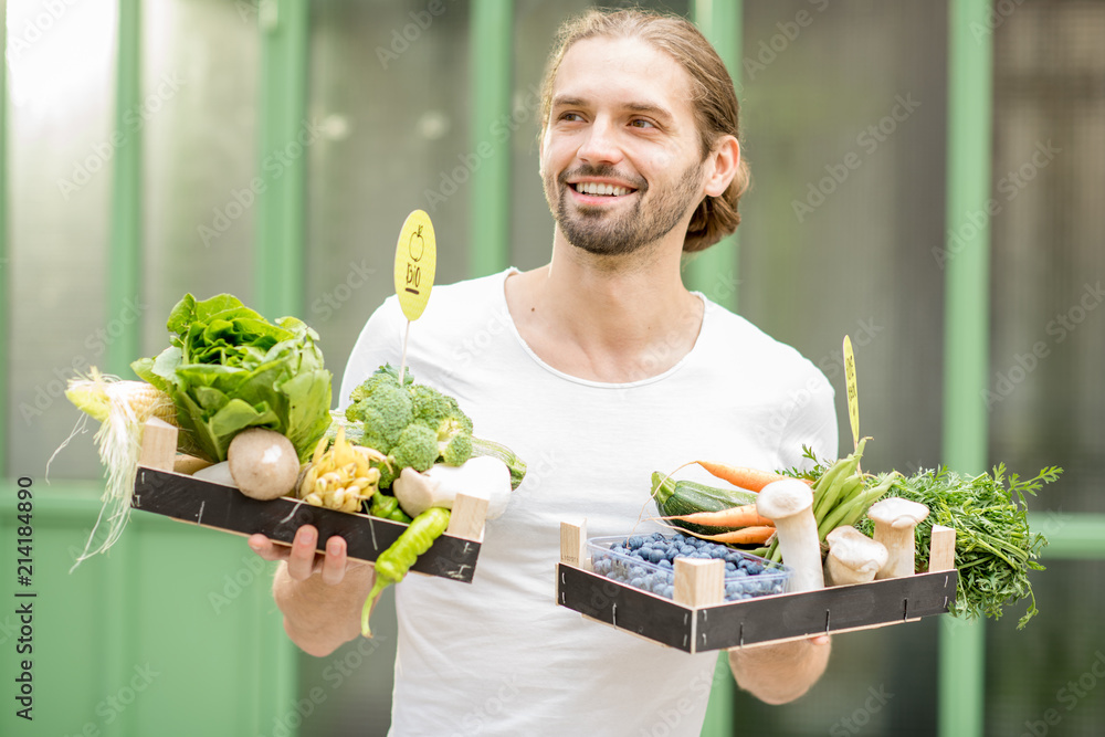 一个英俊男子的画像，他拿着装满新鲜生蔬菜的盒子站在户外的绿色wa上