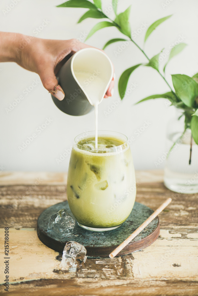 杯子里的冰抹茶拿铁饮料，椰奶由女人的手从水罐里倒出来，白墙