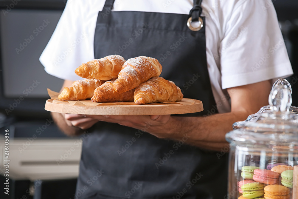 Male baker holding wooden board with delicious croissants in shop