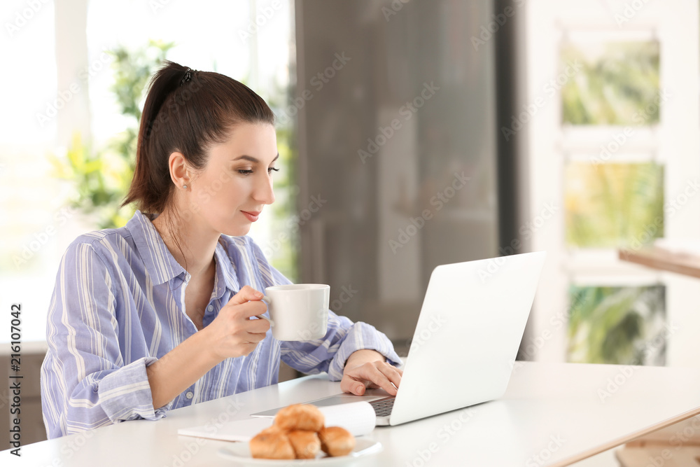 Young female freelancer drinking coffee while working with laptop at home