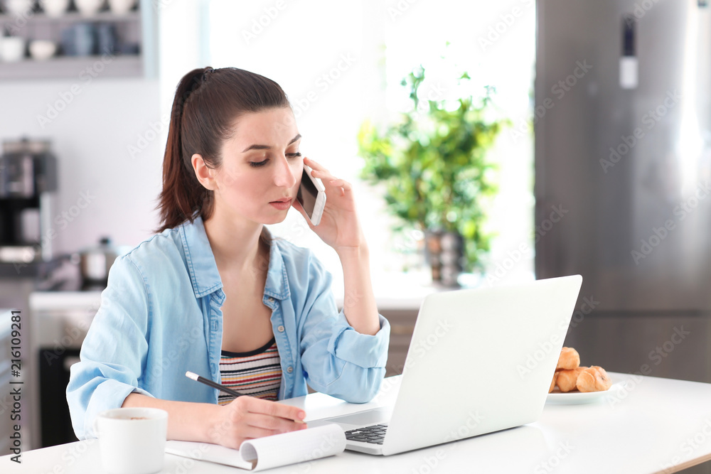 Young female freelancer talking on phone while working with laptop in kitchen