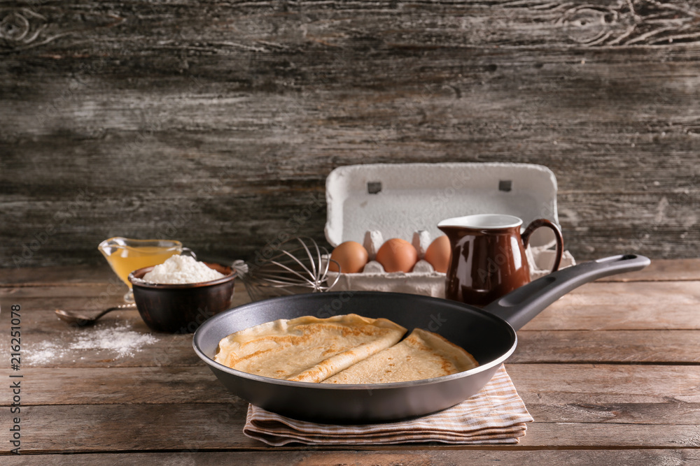 Frying pan with tasty thin pancakes and ingredients on wooden table