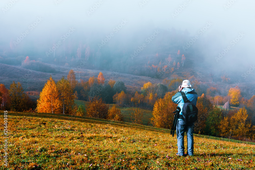 Photographer taking photo of autumn landscape with foggy peaks and orange trees. Ukrainian Carpathia