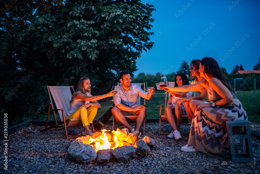 To our great journey! Friends toasting while sitting around firepit.