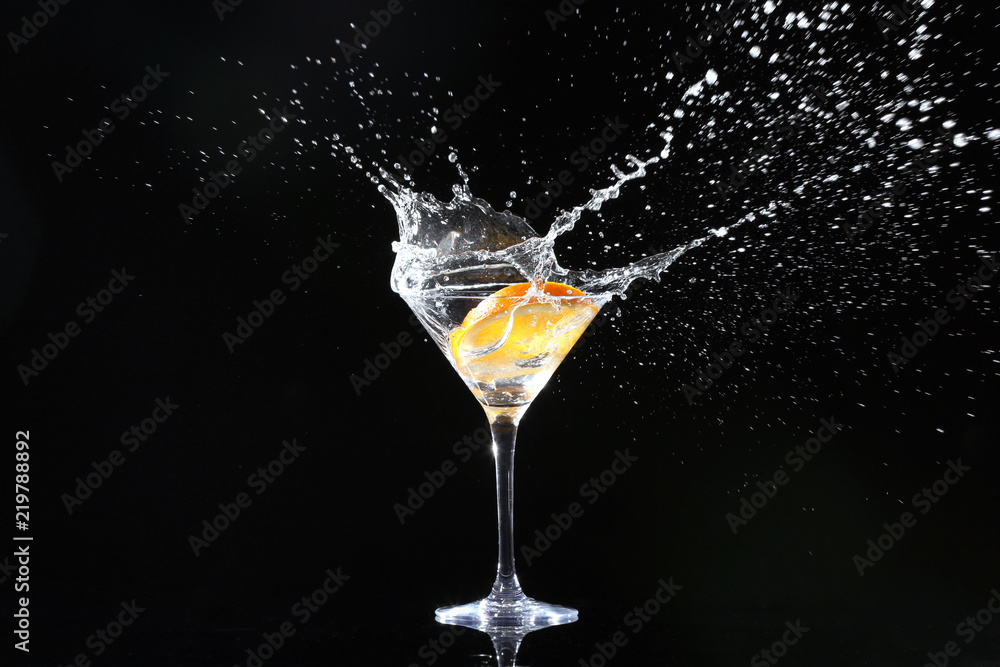 Glass with splashing cocktail and slice of orange on black background