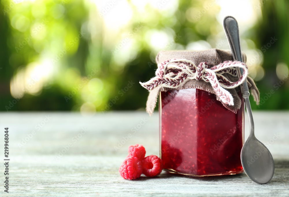 Glass jar with tasty raspberry jam on table against blurred background