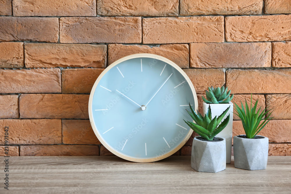 Modern clock with plants on table against brick wall