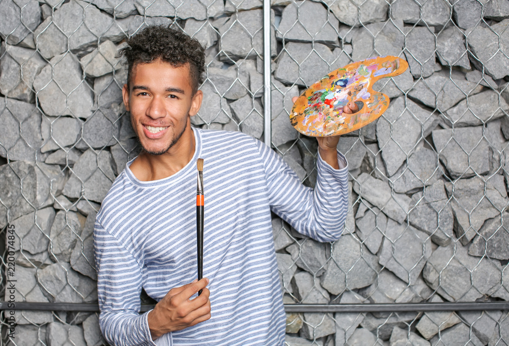 Young African-American artist with palette and brush against stone wall